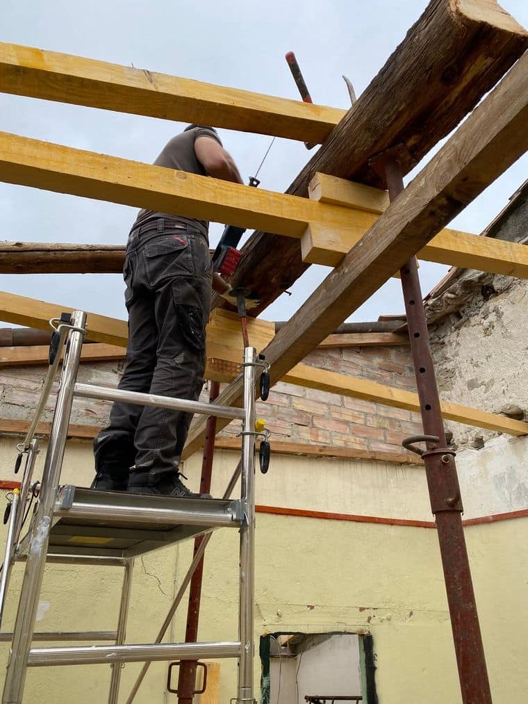 Compagnon sur un échafaudage roulant sous des étais métalliques, pendant la reprise de la structure bois d’une maison ancienne