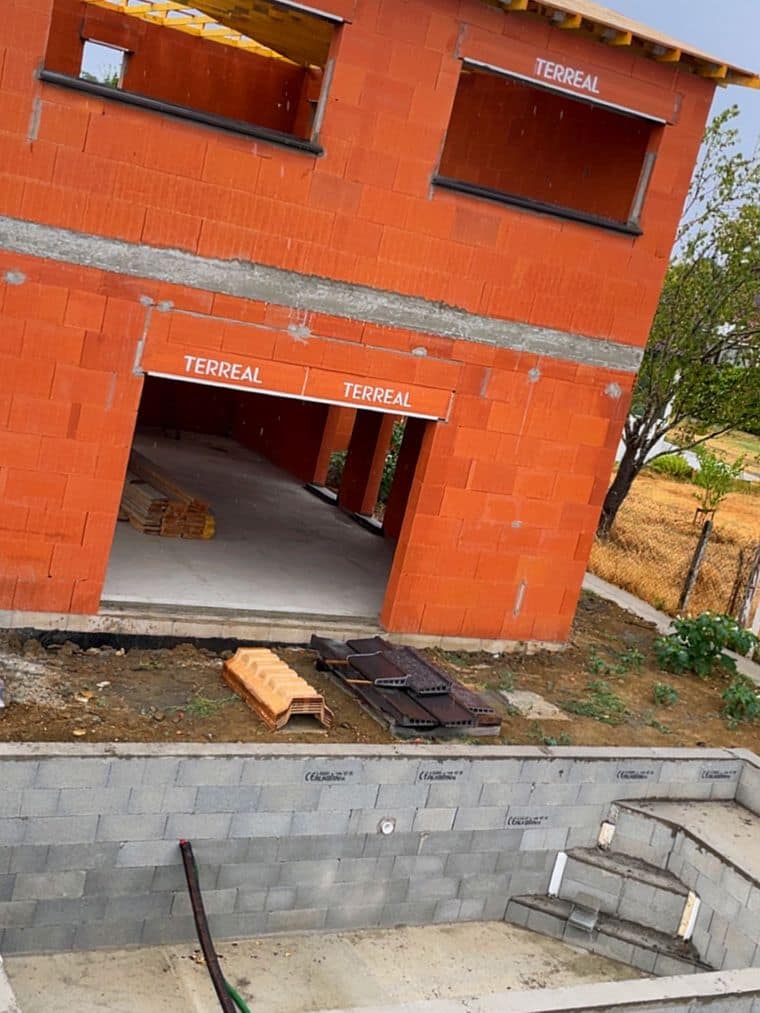 Maison en brique en cours d’élévation sur terrain en pente, murs de soutènement en blocs et emmarchements d’accès au premier plan